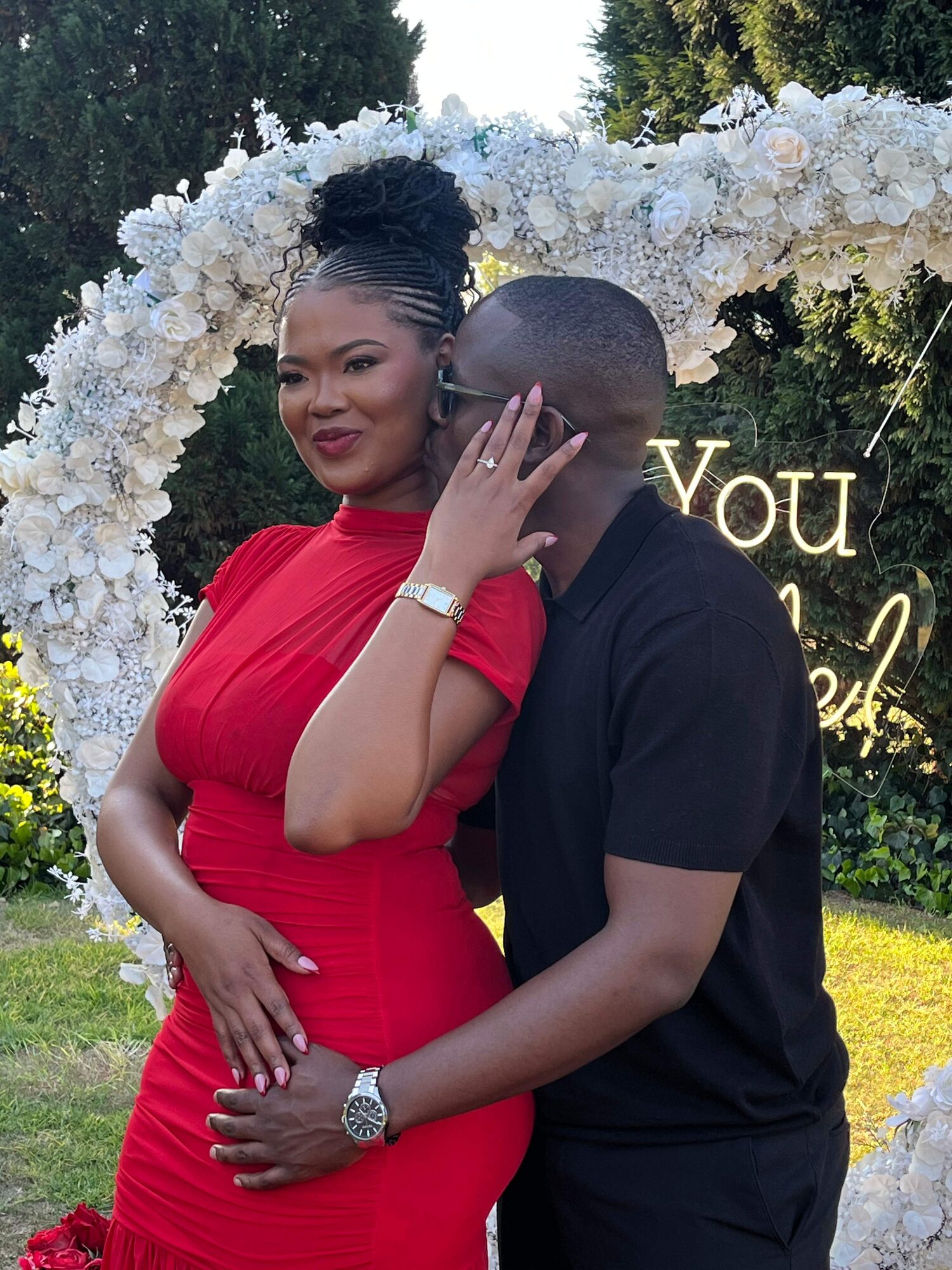 Chukwuma and Ellen embracing beneath a floral arch that reads 'Will You Marry Me?'