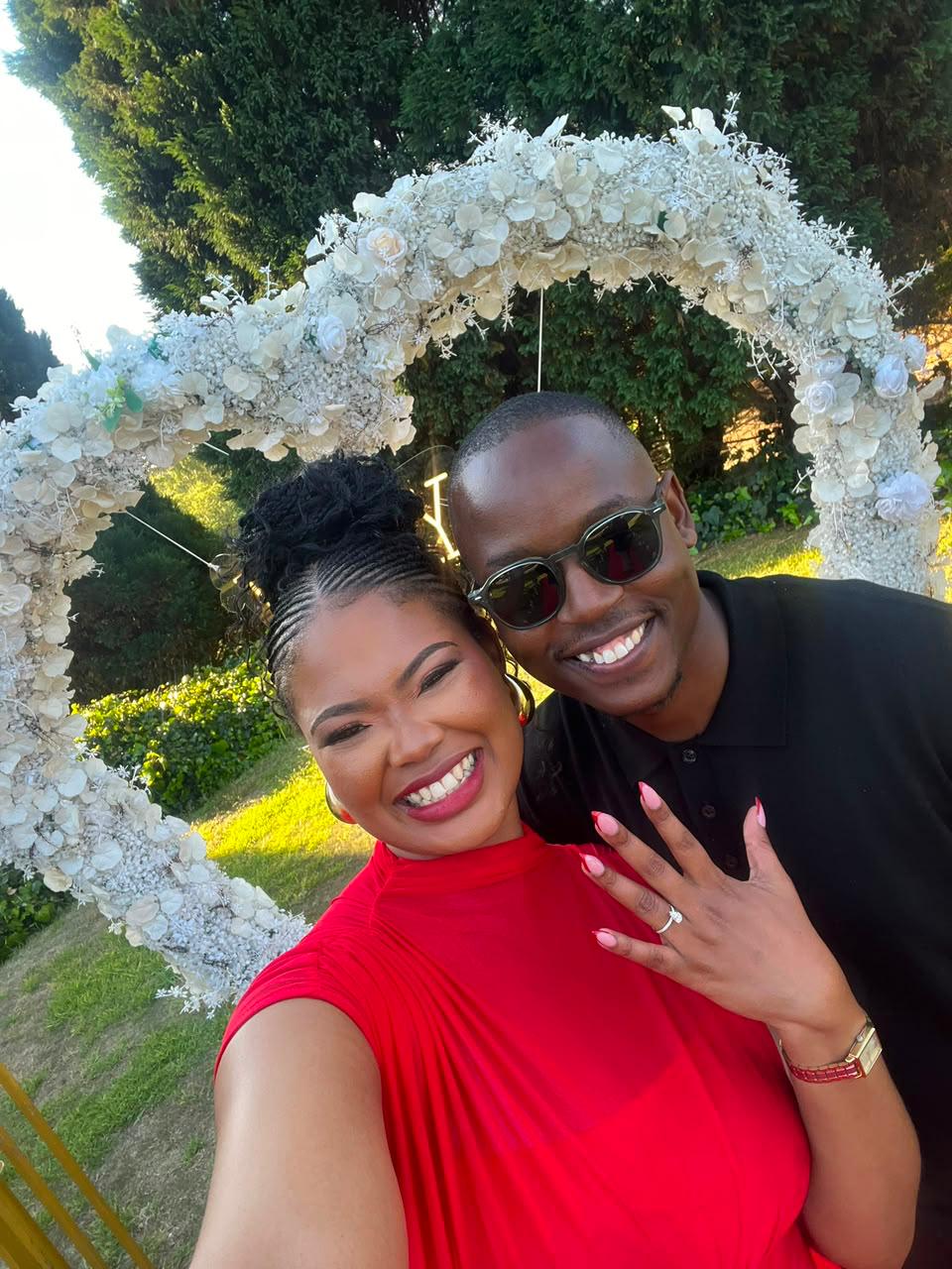 Chukwuma and Ellen smiling together beneath the floral arch, Ellen showing her engagement ring